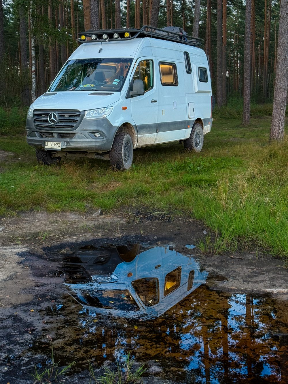 Mercedes Sprinter 4x4 camper reflected in a puddle at dusk in a forest