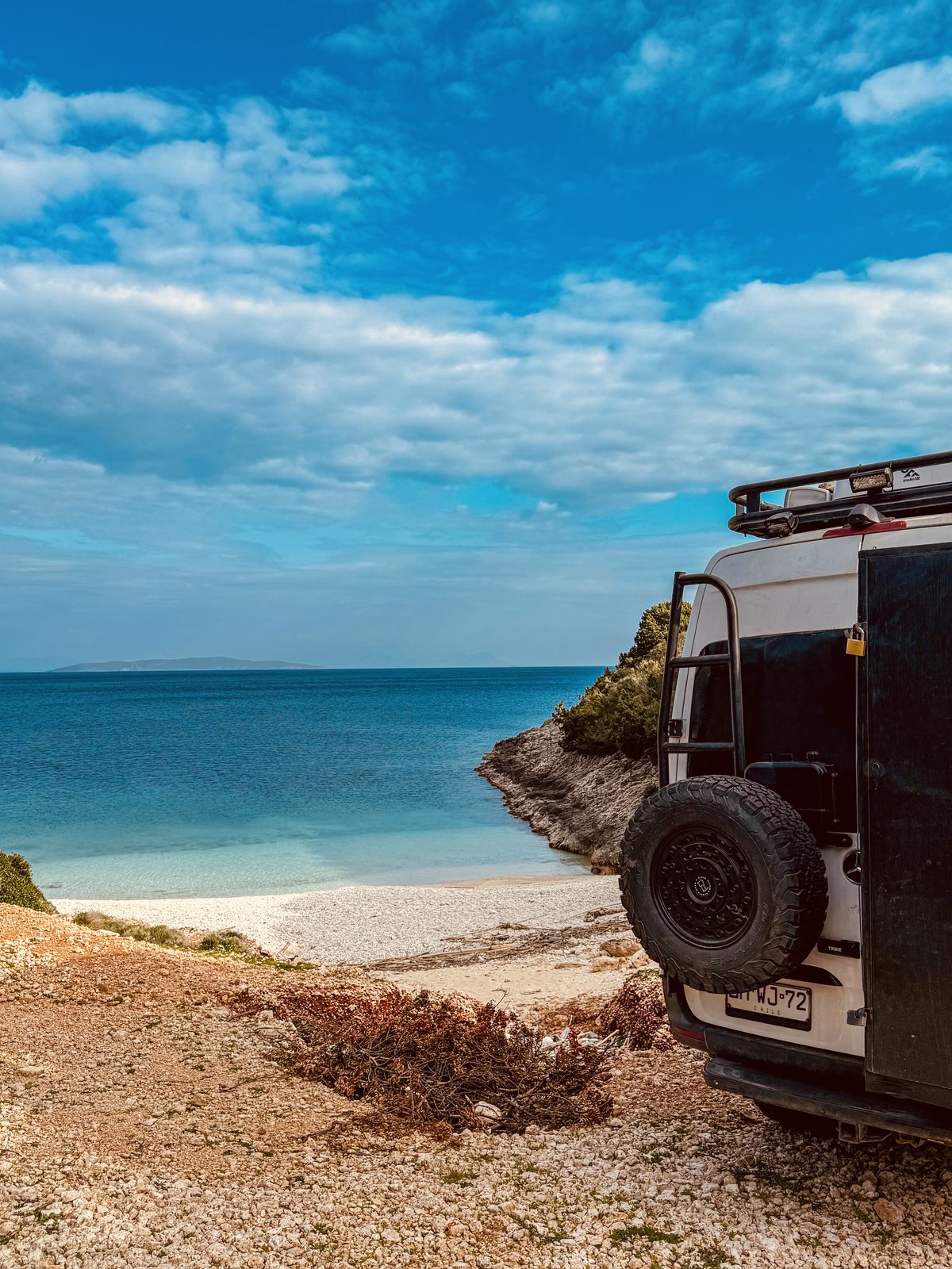 Rear of a Mercedes Sprinter 4x4 camper with spare wheel, overlooking a turquoise cove and pebble beach