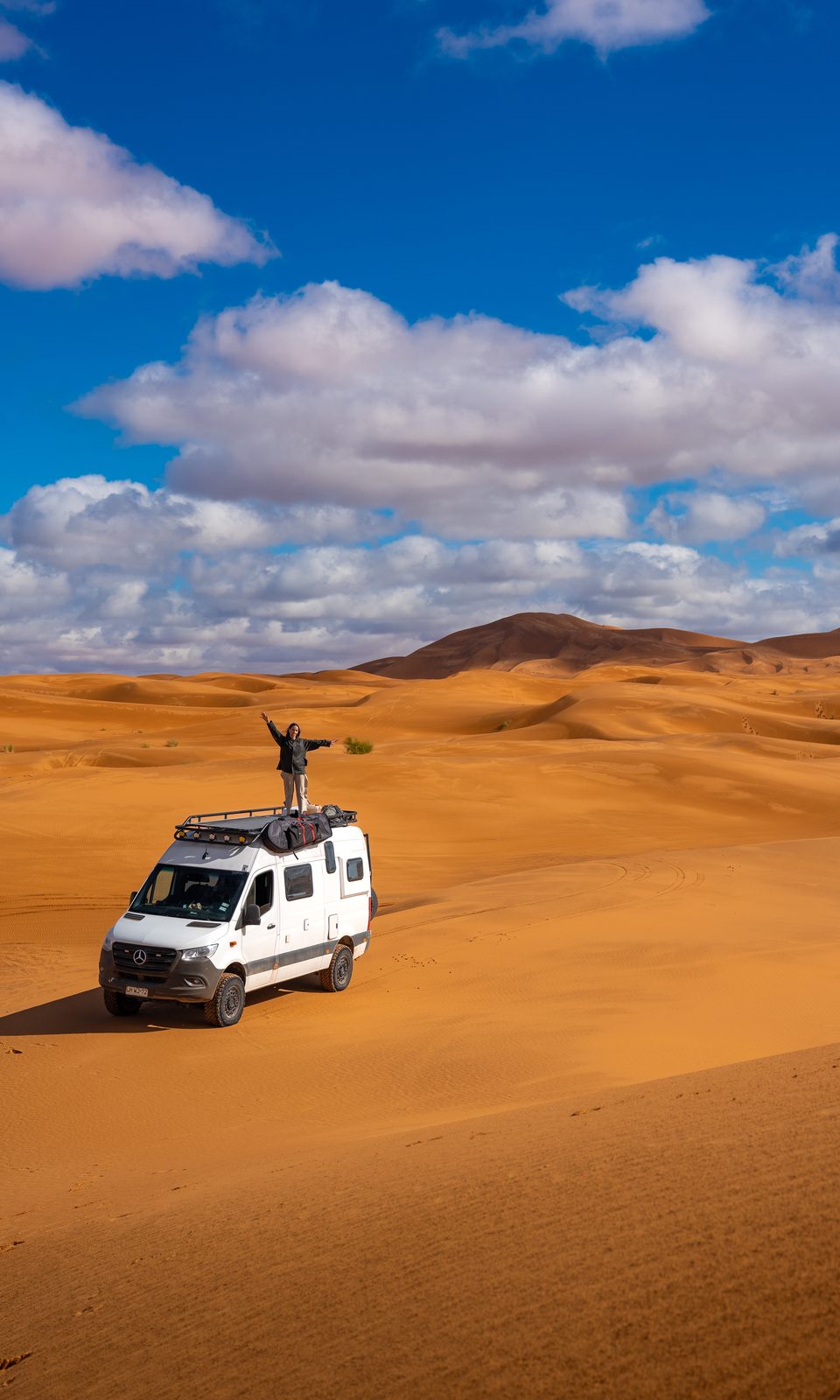 Mercedes Sprinter 4x4 camper crossing orange desert dunes under a blue sky, with a traveler standing on the roof