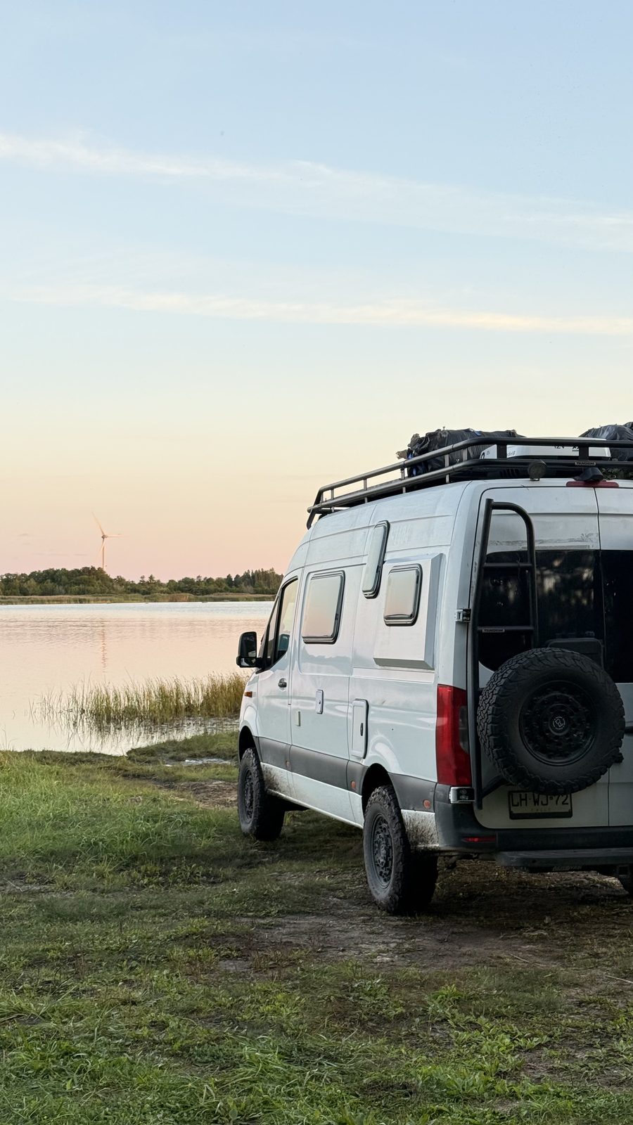 Rear of a Mercedes Sprinter 4x4 camper parked on the grass beside a calm lake at sunset