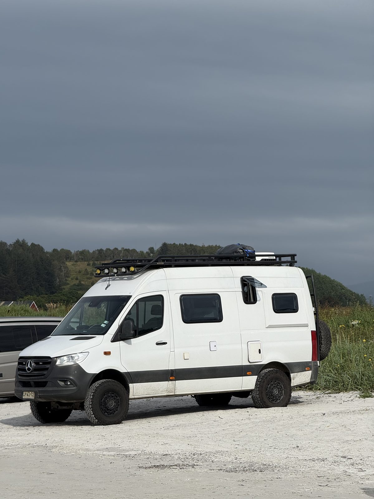 Side view of a Mercedes Sprinter 4x4 camper with roof rack and spare wheel on a pale gravel shore