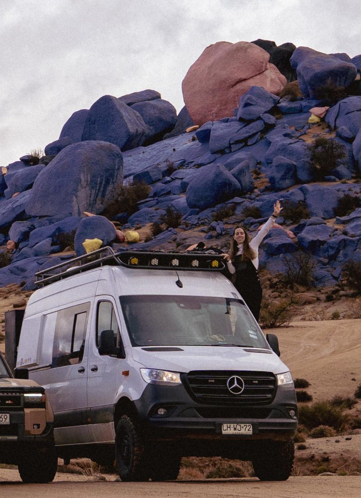 Mercedes Sprinter 4x4 camper in front of a hillside of painted blue boulders, with a traveler waving from the roof